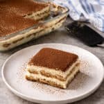 A square slice of tiramisu dusted with cocoa powder sits on a white plate. In the background, an easy tiramisu recipe awaits in a larger glass dish, alongside a serving spatula and a white and blue cloth.