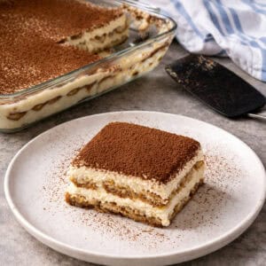 A square slice of tiramisu dusted with cocoa powder sits on a white plate. In the background, an easy tiramisu recipe awaits in a larger glass dish, alongside a serving spatula and a white and blue cloth.