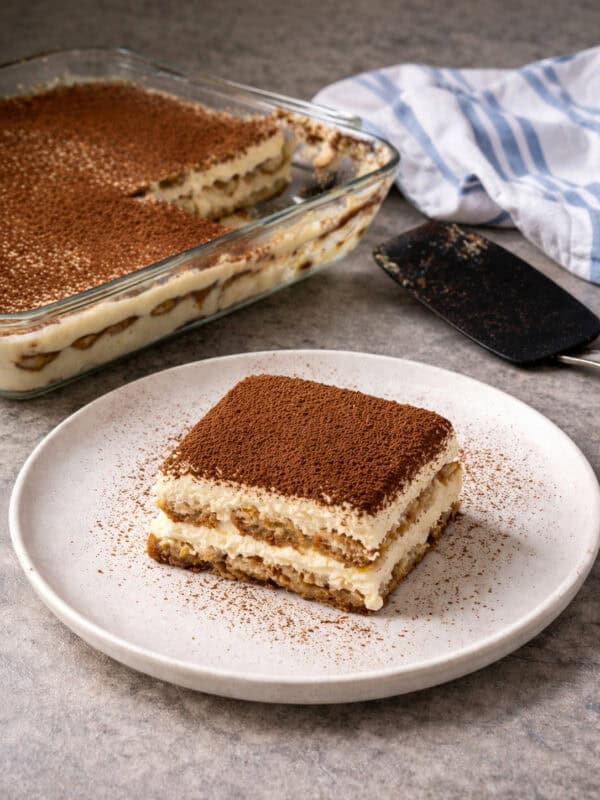 A square slice of tiramisu dusted with cocoa powder sits on a white plate. In the background, an easy tiramisu recipe awaits in a larger glass dish, alongside a serving spatula and a white and blue cloth.