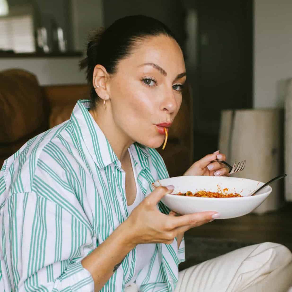A woman with dark hair in a striped shirt is sitting indoors, holding a bowl of pasta and eating a spaghetti noodle while looking at the camera.