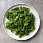 A white plate filled with chopped, cooked flat beans seasoned with herbs and coarse salt, placed on a gray surface—a simple yet flavorful flat bean salad.