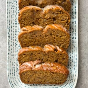 Five slices of Pumpkin Banana Bread are arranged in a row on a rectangular white plate with blue dotted patterns, placed on a light gray surface.