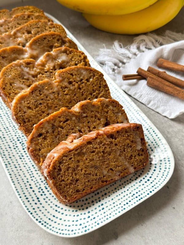 Sliced glazed Pumpkin Banana Bread arranged on a dotted rectangular plate, with bananas, a white cloth, and cinnamon sticks in the background on a light countertop.