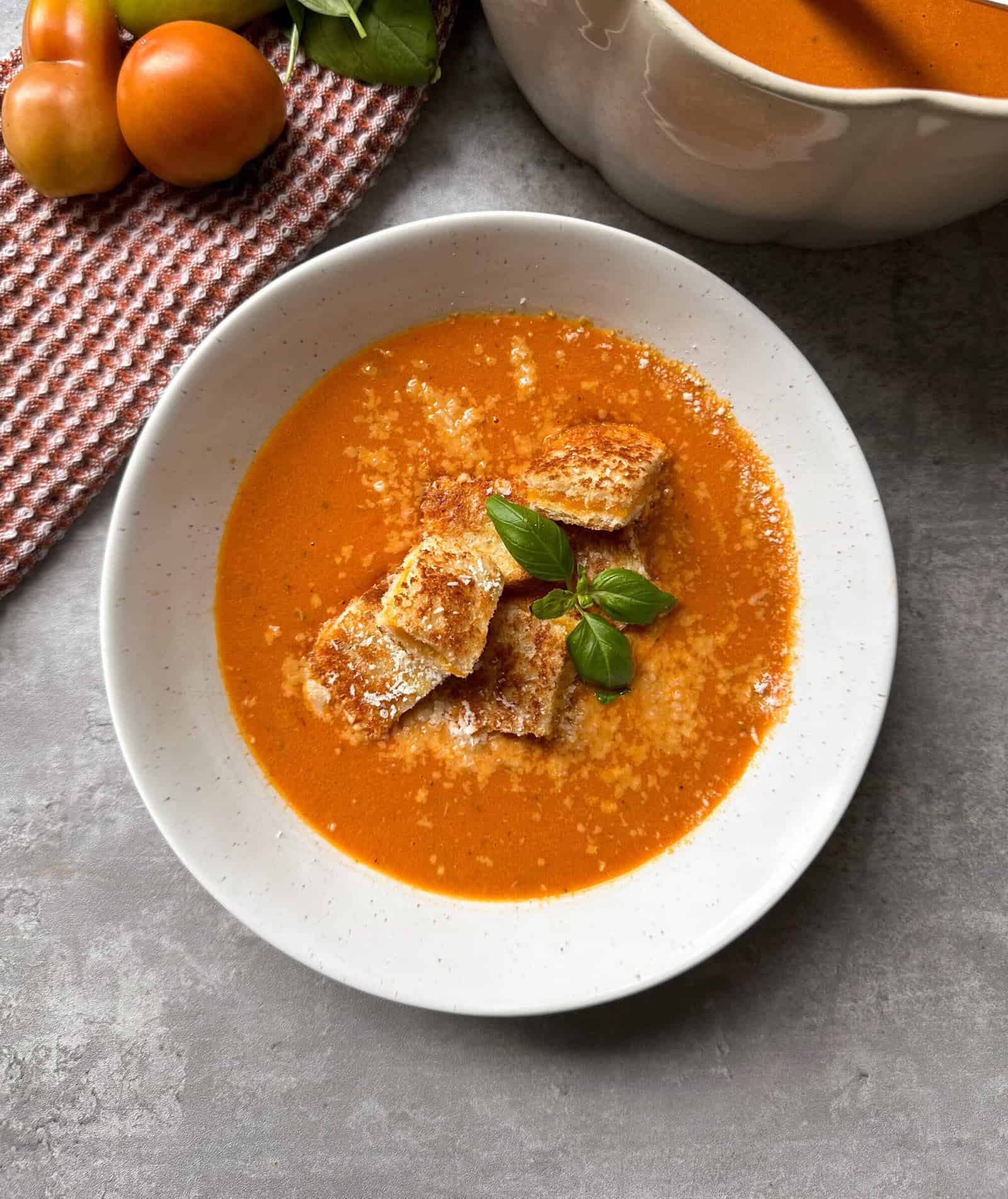 A bowl of creamy roasted tomato soup topped with croutons, grated cheese, and fresh basil sits on a gray surface next to a dish towel, tomatoes, and a pot of soup.