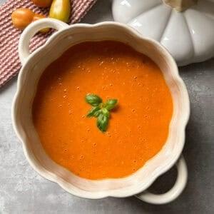 A white, scalloped-edged bowl filled with creamy roasted tomato soup, garnished with fresh basil leaves, sits on a gray surface beside a red dish towel and a few tomatoes.
