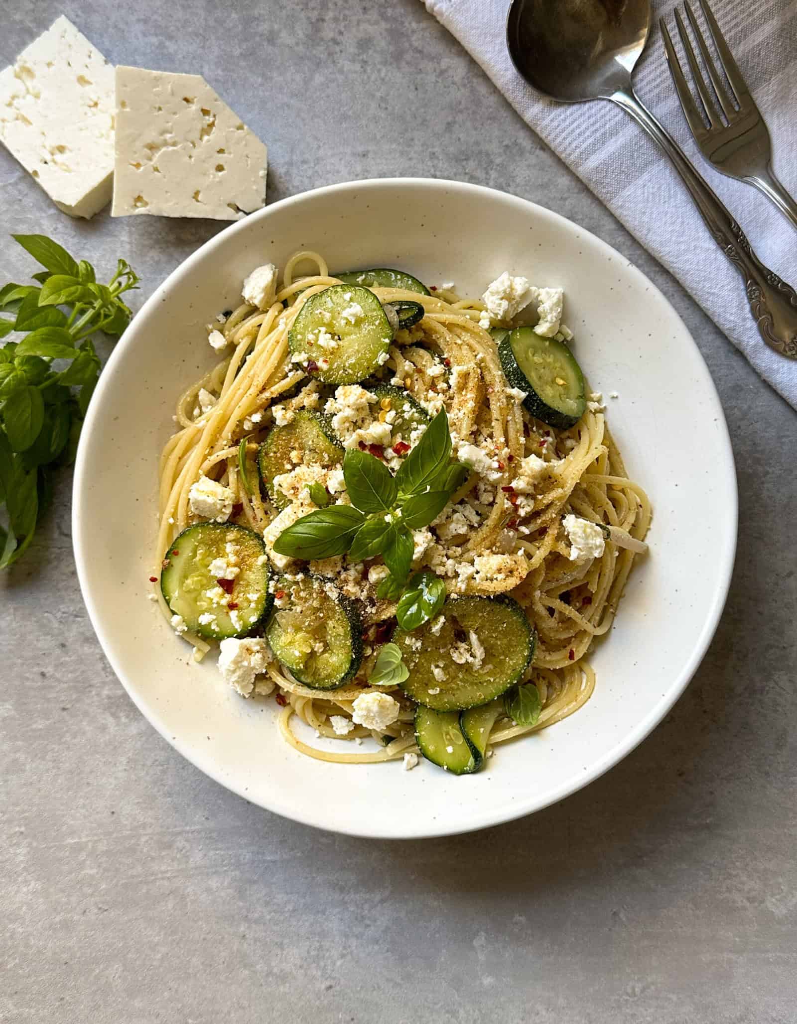 A plate of spaghetti with zucchini and feta is topped with saut&eacute;ed zucchini, crumbled feta, basil leaves, and red pepper flakes. Slices of feta, fresh basil, utensils, and a napkin are nearby on a light gray surface.