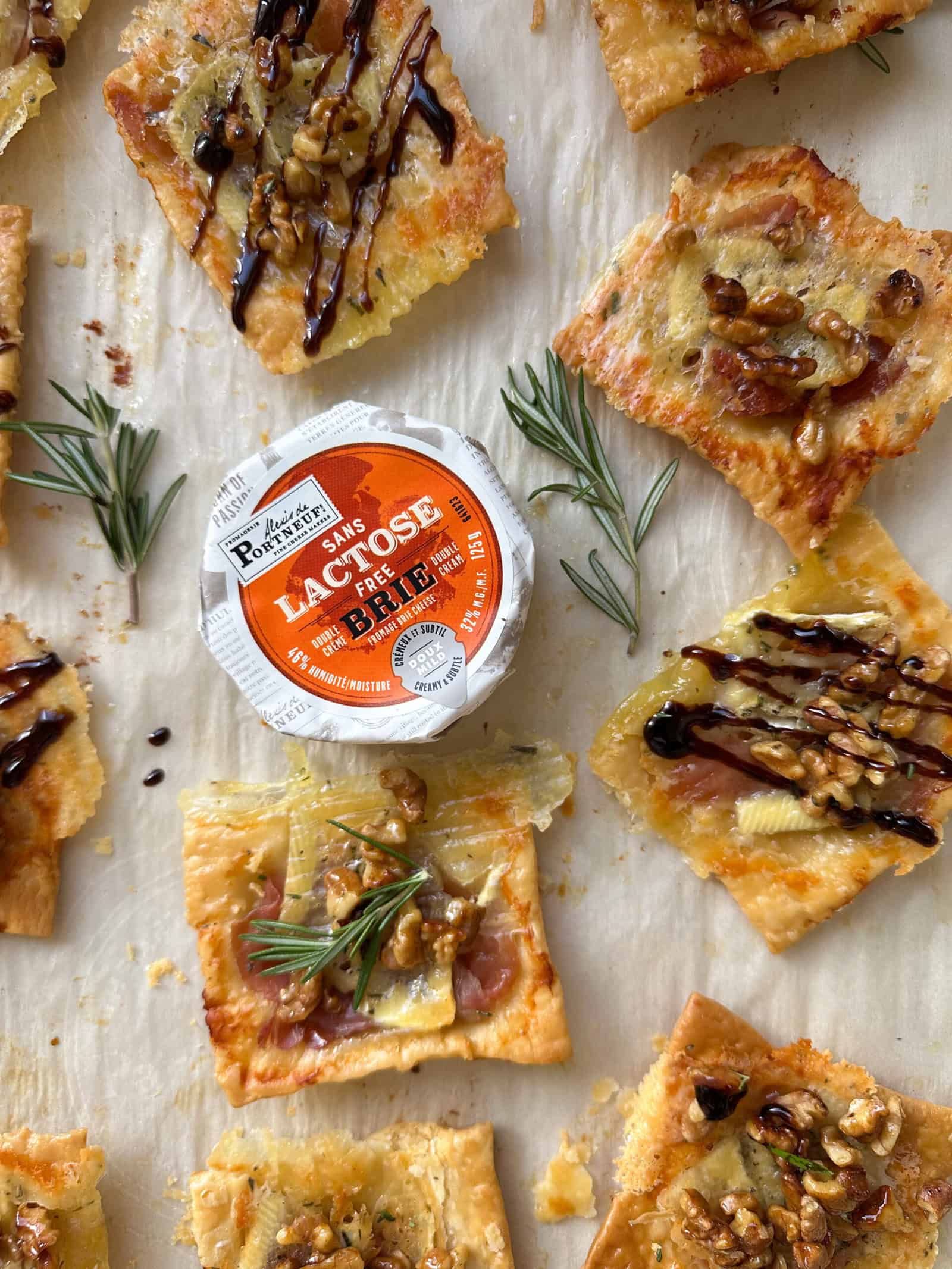 These Puff Pastry Appetizers feature flatbread squares topped with brie, walnuts, and balsamic glaze, arranged around a round container of lactose-free brie; fresh rosemary sprigs complete these festive Holiday Appetizers.