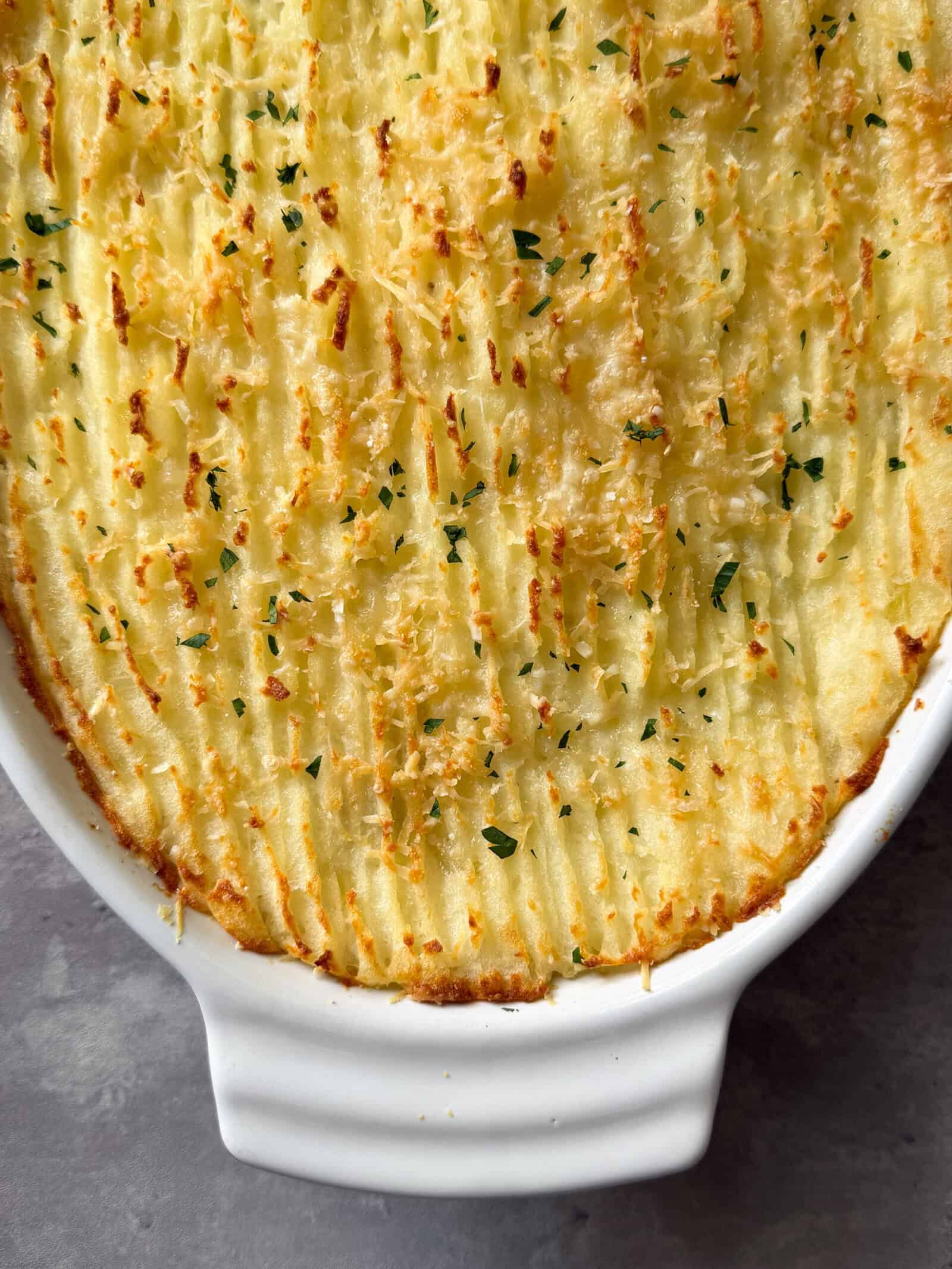 A close-up of a golden-brown baked shepherd's pie, made with an easy cottage pie recipe and topped with mashed potatoes and chopped herbs, in a white ceramic dish.