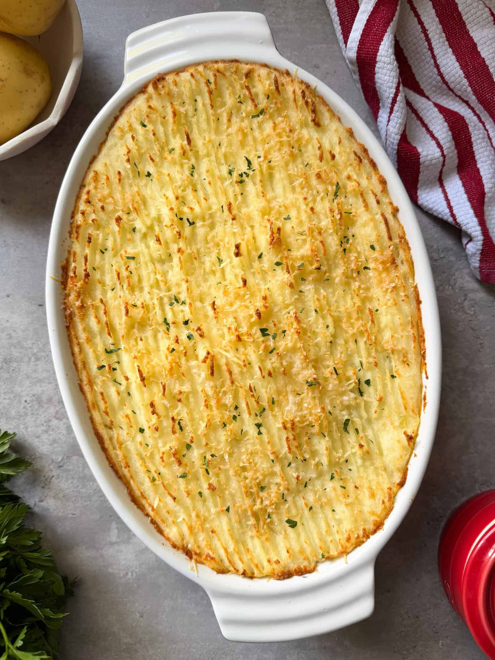Oval white baking dish filled with baked Cottage Pie, topped with golden, textured mashed potatoes and garnished with herbs. A red-striped towel, potatoes, and parsley are visible nearby on a gray surface—perfect for trying an easy Cottage Pie Recipe.