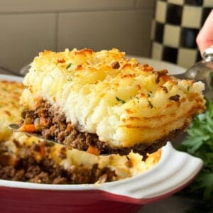 A close-up of a serving of classic Cottage Pie being lifted from a baking dish, showing layers of seasoned meat, vegetables, and golden mashed potatoes. The background includes part of the dish and a checkered container.