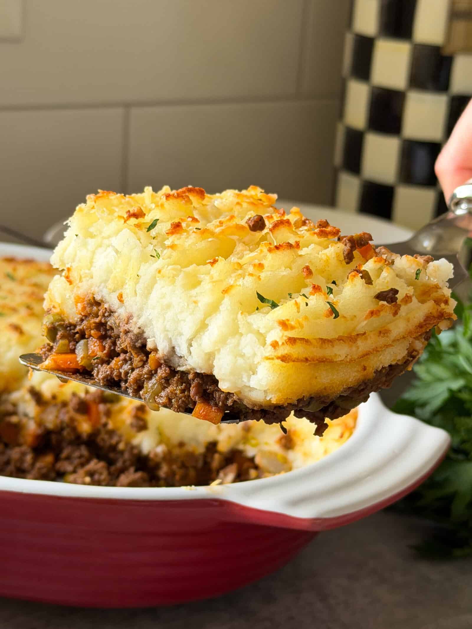 A close-up of a serving of shepherd’s pie, often called cottage pie, showing layers of seasoned ground meat, vegetables, and golden mashed potatoes, being lifted from a red baking dish—perfect for trying an easy Cottage Pie Recipe.
