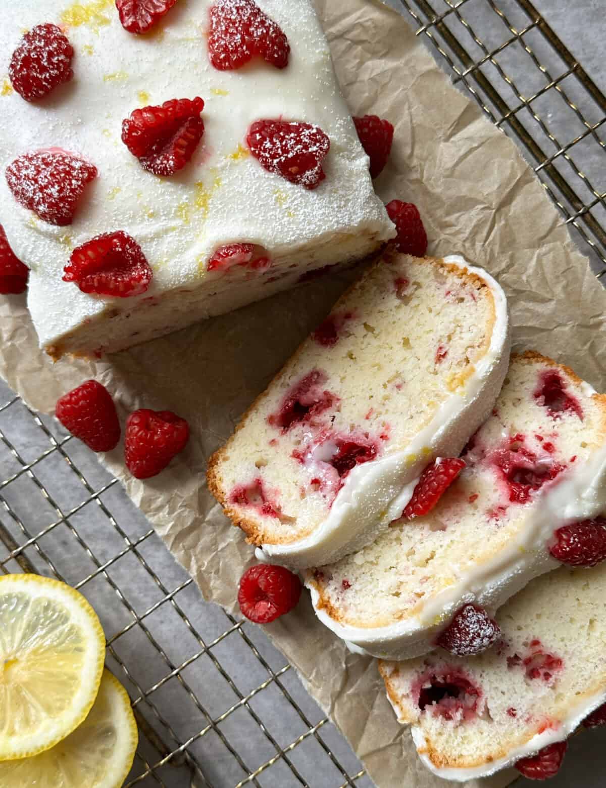 A Raspberry Lemon Loaf recipe shines in this image: a loaf of lemon raspberry cake with white icing, fresh raspberries, and powdered sugar sliced on parchment paper. Extra raspberries and lemon slices rest nearby on a cooling rack.