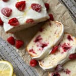 A Raspberry Lemon Loaf recipe shines in this image: a loaf of lemon raspberry cake with white icing, fresh raspberries, and powdered sugar sliced on parchment paper. Extra raspberries and lemon slices rest nearby on a cooling rack.