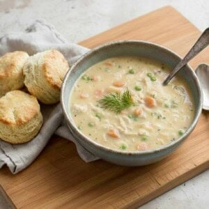 A bowl of creamy chicken pot pie soup with peas and carrots, garnished with dill, sits on a wooden board next to two flaky biscuits and a silver spoon.
