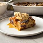 A slice of Blueberry French Toast Casserole with visible berries, topped with whipped cream and syrup, served on a white plate with a baking dish in the background. Enjoy this delightful Blueberry French Toast Casserole Recipe for breakfast or brunch!.