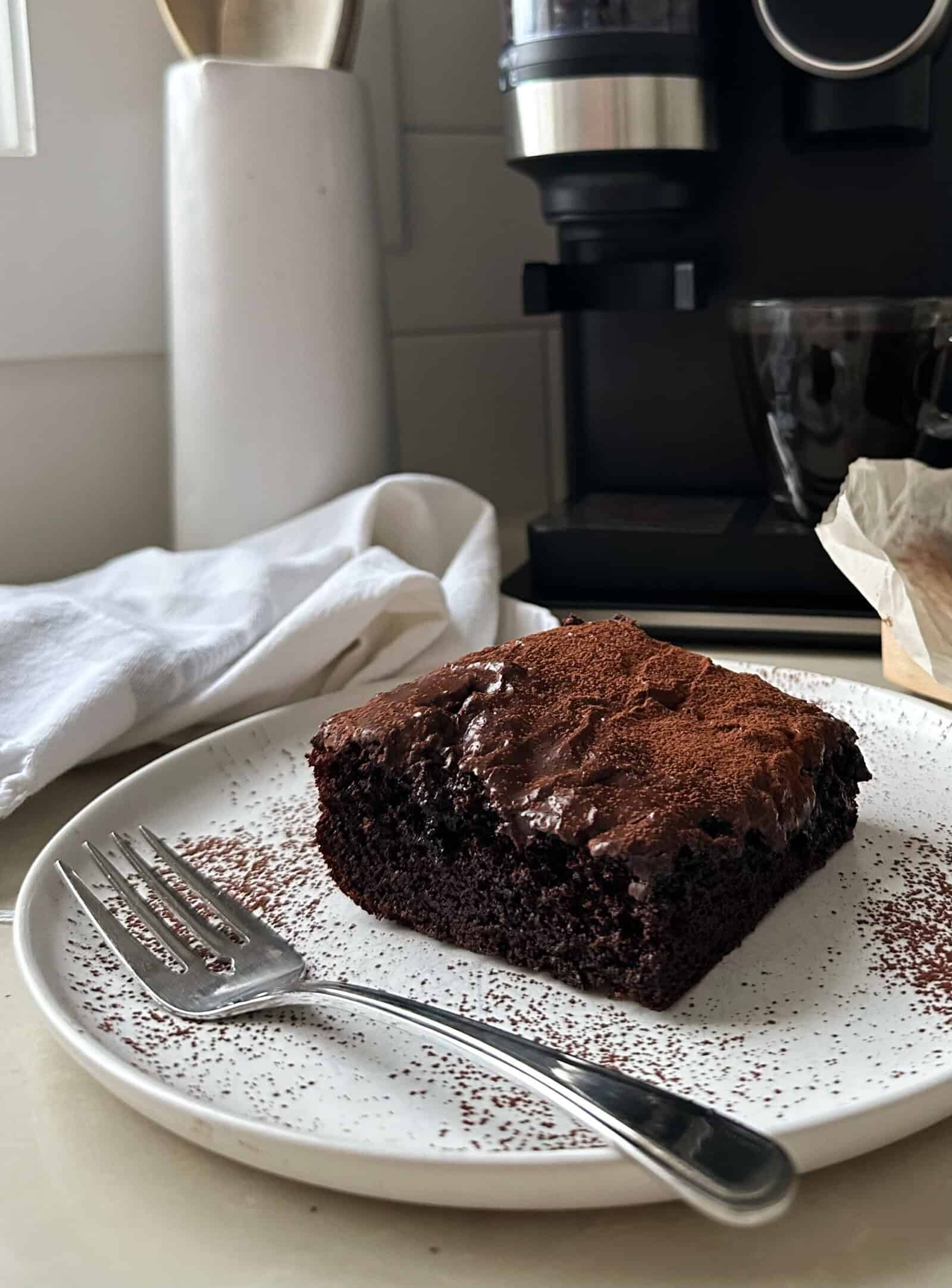 A piece of chocolate sheet cake with icing sits on a white plate with a fork beside it. A coffee maker, cup, and kitchen utensils are in the background, and a white cloth is draped on the counter.