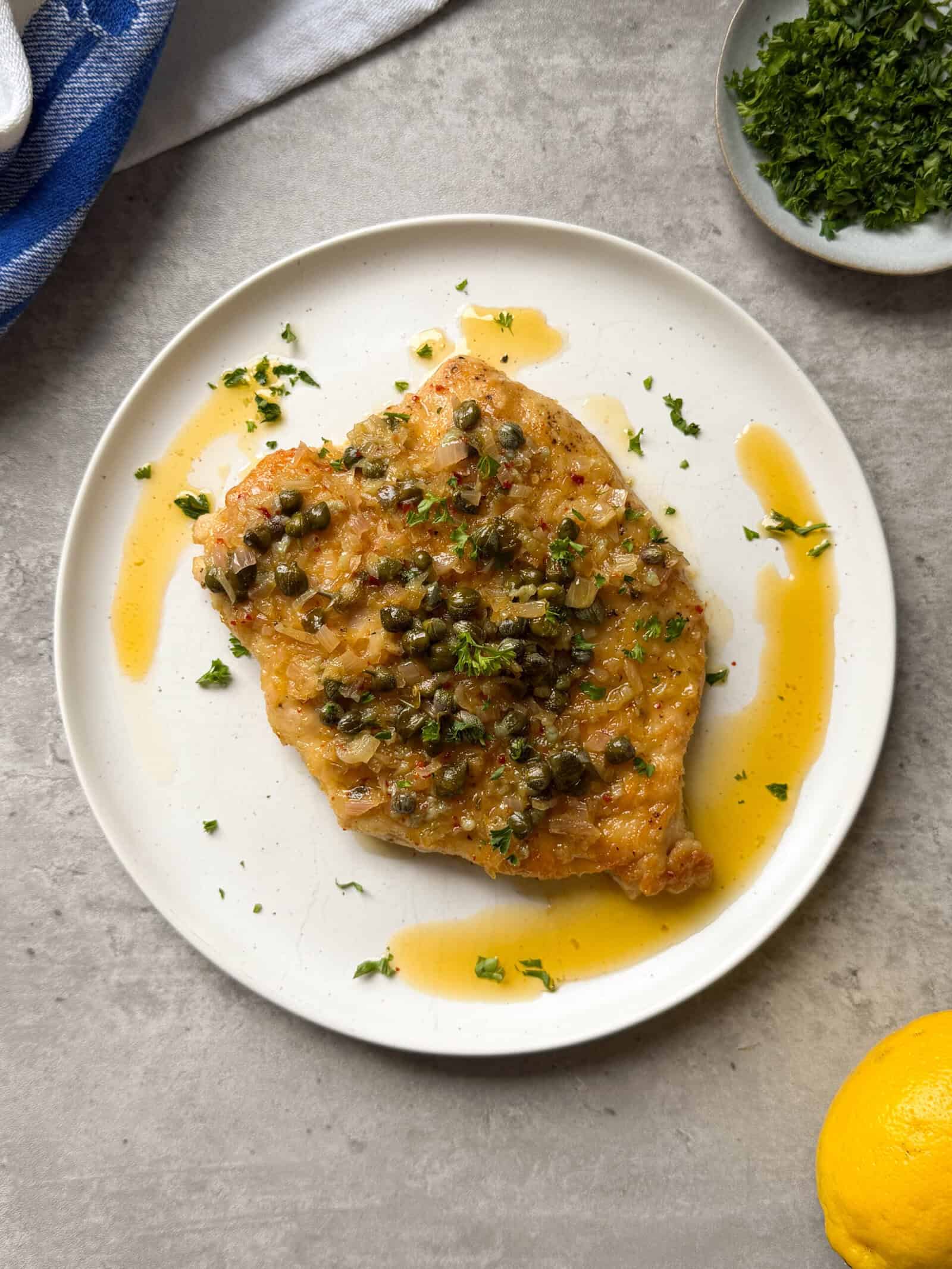 A cooked chicken breast topped with capers and chopped herbs sits on a white plate with a pool of sauce&mdash;an inviting display of easy chicken piccata recipe essentials. A bowl of parsley, a lemon, and a blue towel rest nearby on a light surface.
