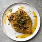 A cooked chicken breast topped with capers, chopped herbs, and a light sauce, served on a round white plate over a gray stone surface—perfect for anyone seeking the best chicken piccata recipe.