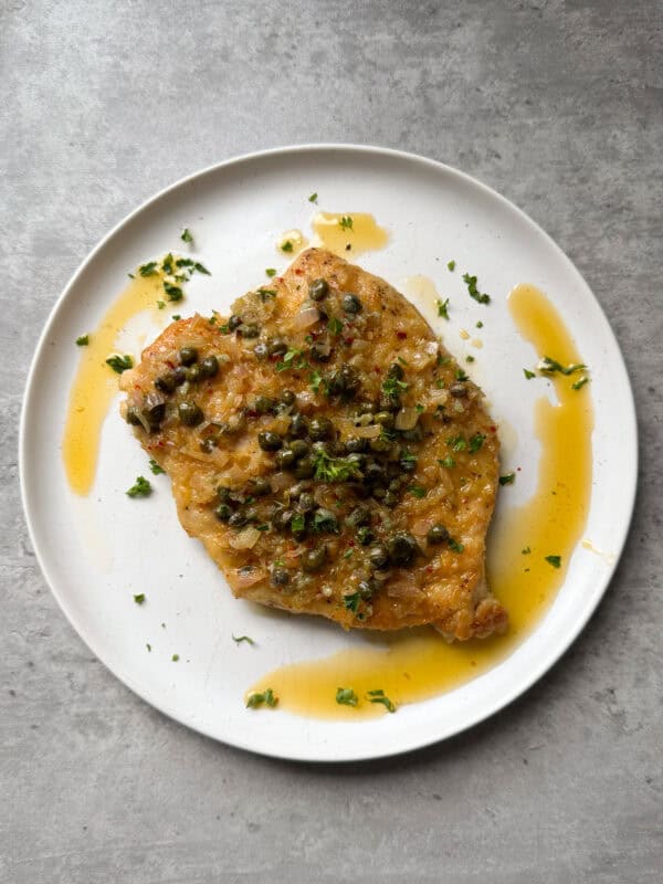 A cooked chicken breast topped with capers, chopped herbs, and a light sauce, served on a round white plate over a gray stone surface—perfect for anyone seeking the best chicken piccata recipe.