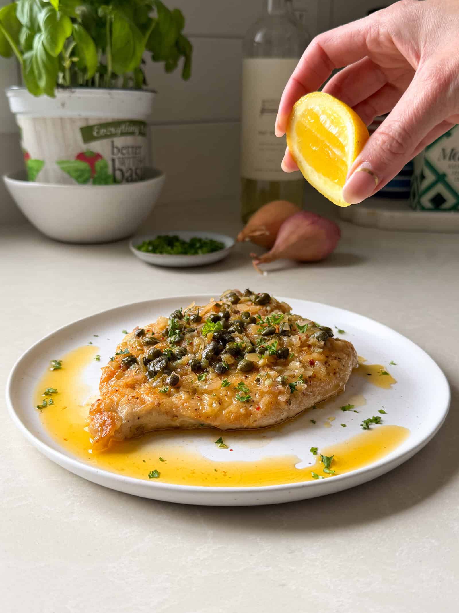A hand squeezes a lemon wedge over a plate of easy chicken piccata recipe, garnished with capers and parsley, with sauce pooling on the plate; kitchen items are blurred in the background.