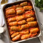 A baking dish filled with neatly arranged cabbage rolls recipe in tomato sauce, baked to a golden brown. Parsley, a serving fork, and a napkin are nearby on a light gray surface.