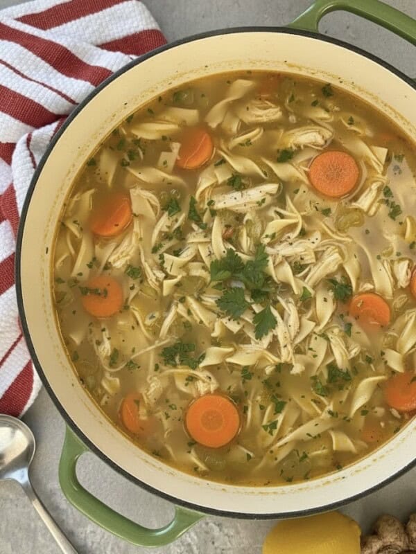 A pot of Easy Chicken Noodle Soup with shredded chicken, sliced carrots, noodles, and parsley in a savory broth. A red and white striped towel and spoon sit beside the pot for cozy serving.