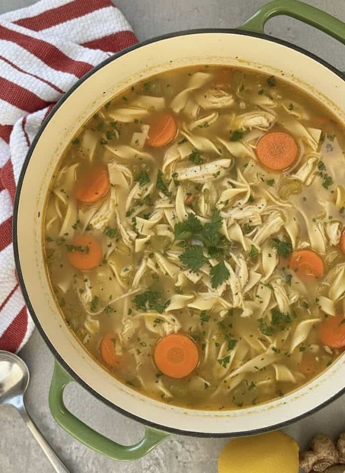 A pot of Easy Chicken Noodle Soup with shredded chicken, sliced carrots, noodles, and parsley in a savory broth. A red and white striped towel and spoon sit beside the pot for cozy serving.