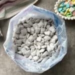 A bag filled with Chex Mix Puppy Chow, those irresistible powdered sugar-covered snacks, sits on a gray surface. Next to it is a white dish holding colorful heart-shaped candies, adding a playful touch. A striped cloth is partially visible in the corner.