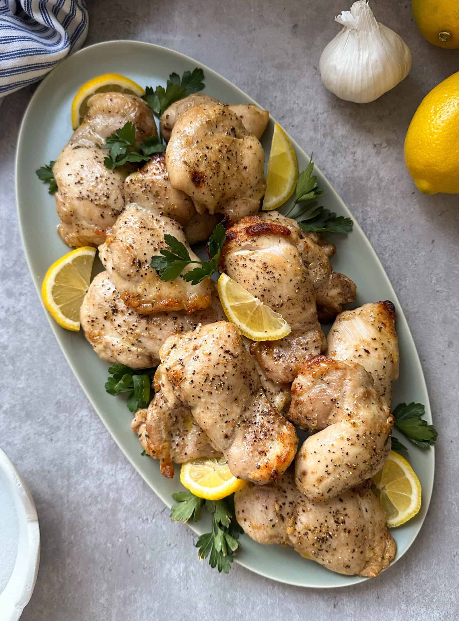 A platter of seasoned baked chicken thighs, enhanced by the Best Chicken Marinade Recipe, is garnished with lemon slices and fresh parsley. The dish is accompanied by bulbs of garlic and whole lemons on a gray surface.