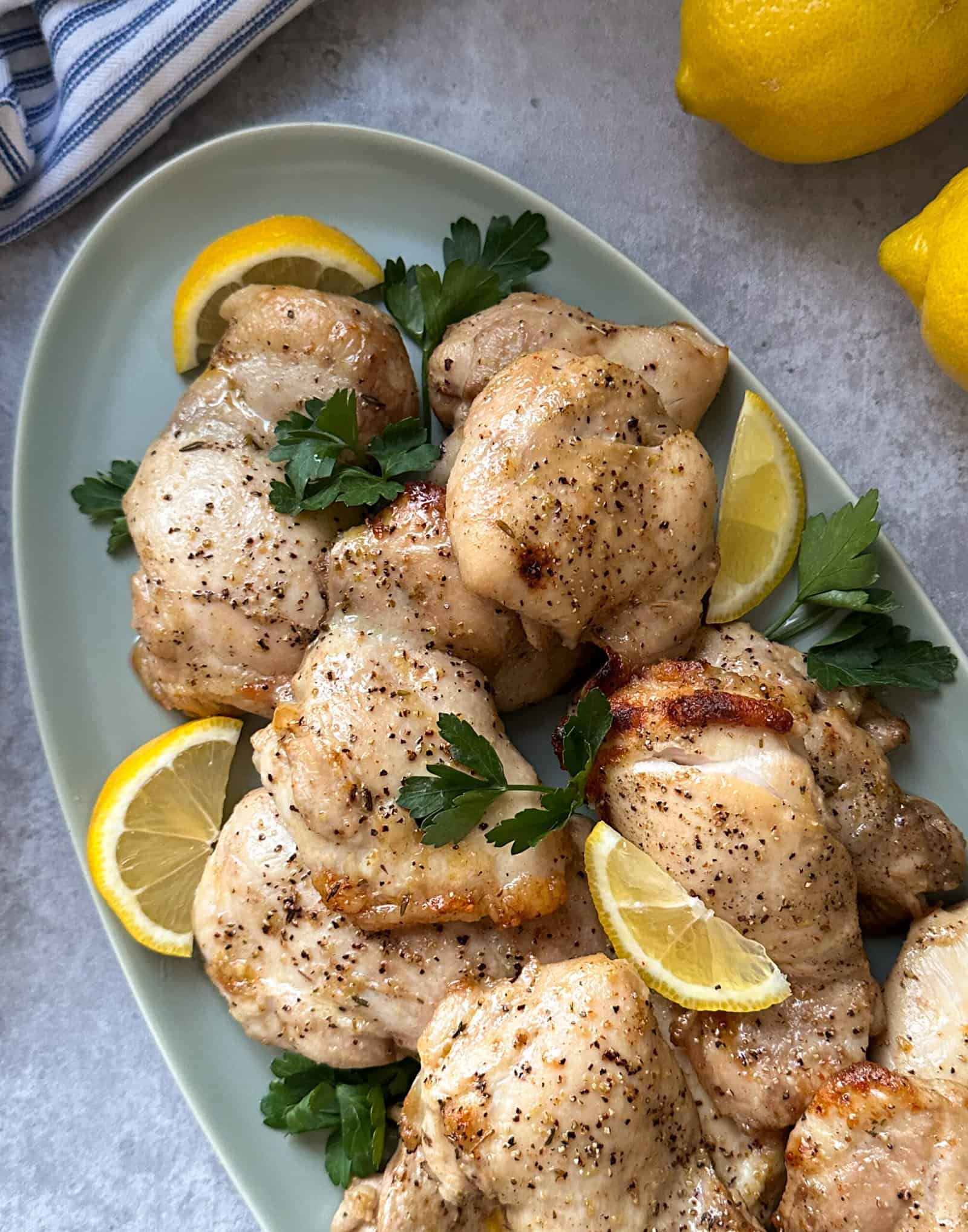 Grilled chicken pieces, marinated to perfection using the best chicken marinade recipe, are garnished with lemon slices and parsley on an oval platter. The dish is artfully surrounded by whole lemons and a striped cloth, set on a light gray surface.