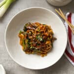 A delightful bowl of noodles featuring a beef and broccoli stir fry, adorned with greens, sesame seeds, and sauce on a gray surface. Nearby lie fresh green onions, ginger, chopsticks, and a red-striped napkin.