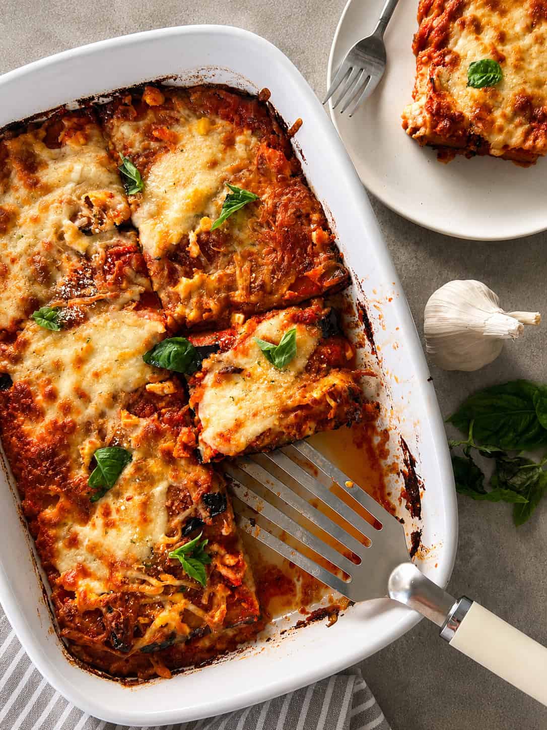 A baked Eggplant Parmigiana in a white dish, partially served, with a serving spatula. A slice sits on a plate nearby. Fresh basil and garlic are beside the dish, and the parmigiana di melanzane is topped with melted cheese and sauce.
