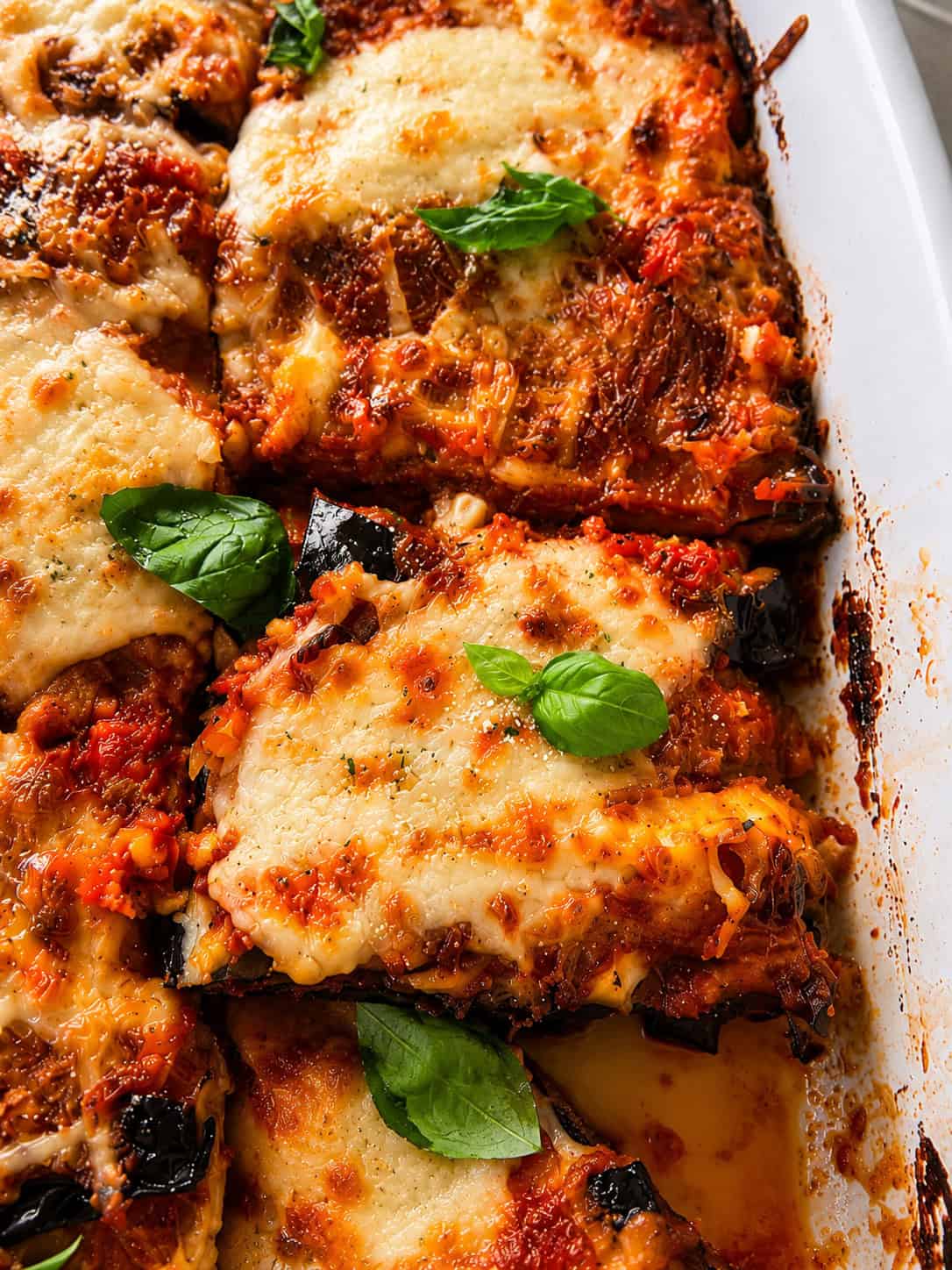 Close-up of baked Eggplant Parmigiana in a white dish, topped with golden melted cheese, tomato sauce, grated parmesan, and fresh basil leaves. One serving is partially lifted, showing the delicious layers and crispy edges of this classic Eggplant Parmigiana Recipe.