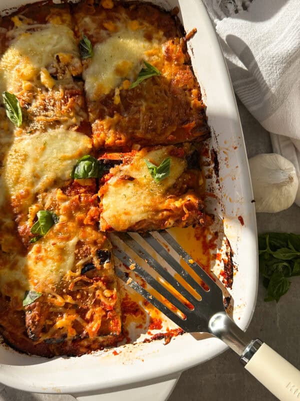 A baked dish of cheesy lasagna with tomato sauce and basil leaves in a white rectangular baking dish, reminiscent of an Eggplant Parmigiana. A slice is being lifted with a spatula, and a garlic bulb and basil leaves are nearby on a gray surface.