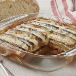 A glass baking dish with baked Eggplant Parmigiana, topped with melted cheese and herbs, sits on a countertop next to a slice of rustic bread, a fork, and a red-and-white striped napkin.