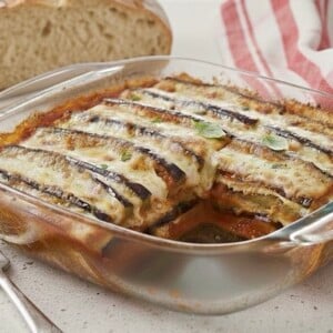 A glass baking dish with baked Eggplant Parmigiana, topped with melted cheese and herbs, sits on a countertop next to a slice of rustic bread, a fork, and a red-and-white striped napkin.