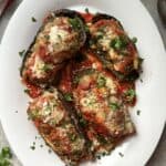 Four halves of baked stuffed eggplant topped with tomato sauce and melted cheese are garnished with chopped herbs on a white plate. Perfect for your next stuffed eggplant recipe, this dish is set against a backdrop of a red and white towel with scattered herbs.