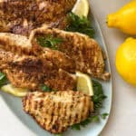 Succulent grilled chicken breasts, infused with the zest of a yogurt marinade recipe, are garnished with parsley and lemon wedges on a blue plate. Two whole lemons rest beside it on a light-colored surface for an extra touch of freshness.