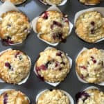 A baking tray filled with twelve blueberry lemon muffins in white paper wrappers. They are golden brown, showcasing hints of baked blueberries and a zesty twist, topped with a crumbly texture.