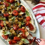 A white platter features a Boursin and Roasted Veggie Spread topped with roasted cherry tomatoes, eggplant, and fresh basil. A hand holds a fork, eager to taste. Two eggplants and a red-striped kitchen towel complete the appetizing scene in the background.
