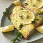 Two pieces of Lemon Dill Salmon topped with a creamy herb sauce and lemon slices are served on a green plate. Garnished with fresh dill and parsley, this delightful dish is accompanied by lemon wedges for an extra citrusy kick.