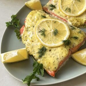 Two salmon fillets topped with a creamy sauce and lemon slices are artfully presented on an oval plate. This delectable dish, a classic Lemon Dill Salmon recipe, is garnished with fresh dill and parsley, accompanied by zesty lemon wedges on the side.