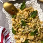 A decorative white plate holds a serving of orecchiette pasta in a creamy sauce, complemented by asparagus, peas, and grated cheese. Fresh basil leaves add a hint of green. A gold serving spoon rests nearby on a red and white striped towel.