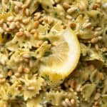 Close-up of Lemon Herb Pasta Salad featuring bowtie pasta tossed in pesto, garnished with toasted pine nuts and a slice of lemon. This easy pasta salad is coated in a green herb mixture, with the lemon adding a pop of yellow.