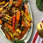 A platter of Honey Roasted Carrots garnished with herbs and crispy garlic slices, served on a bed of creamy spread. A gold serving utensil and a red-and-white striped towel are beside the plate.
