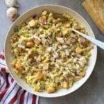 A bowl of Caesar Pasta Salad with rotini, croutons, diced ham, lettuce, and creamy dressing sits beside a striped red and white towel, whole garlic bulbs, and a baguette on a gray surface. Try this Caesar Pasta Salad Recipe for your next meal!.