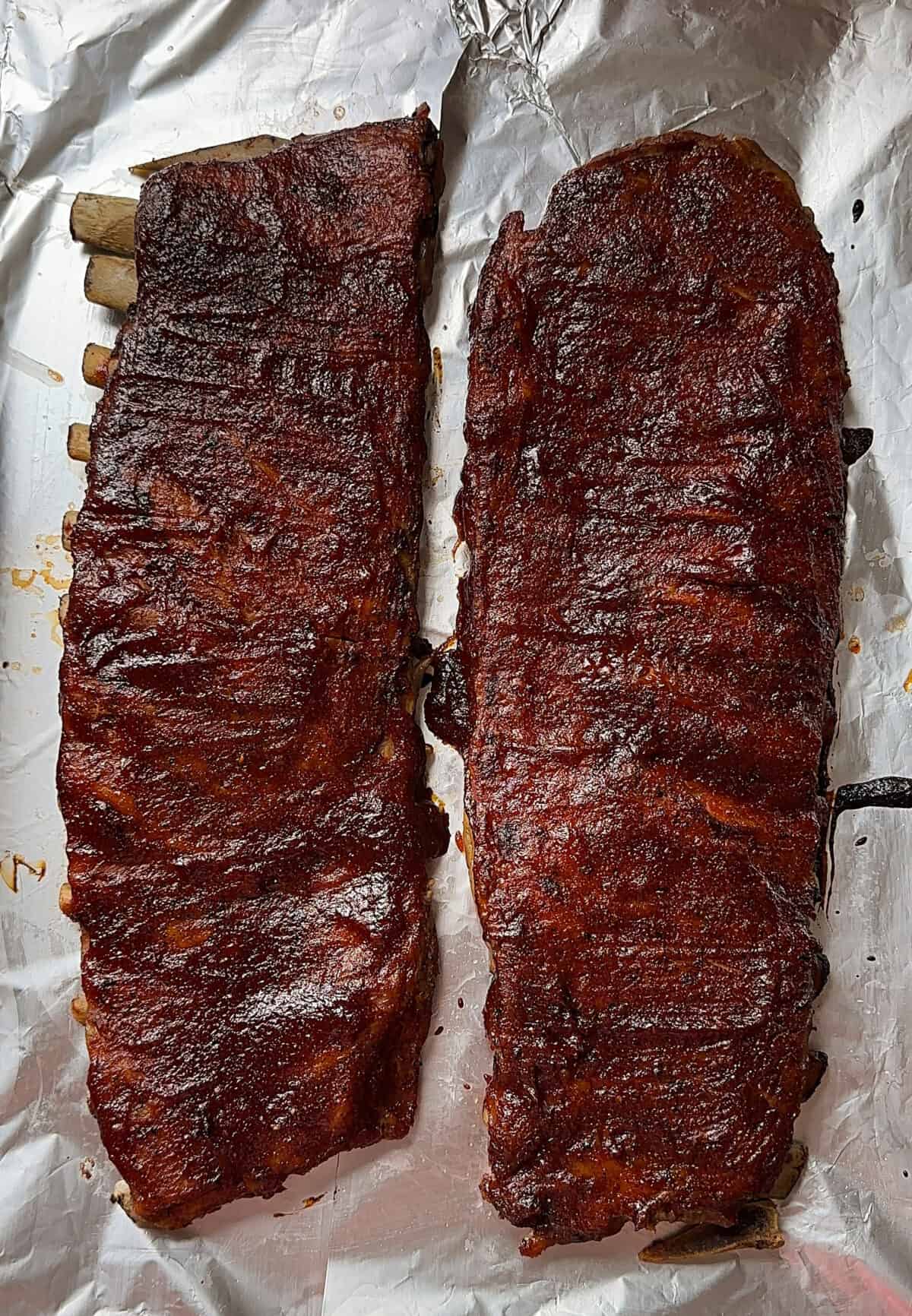 Two racks of oven baked bbq ribs with a dark, glossy sauce rest on crinkled aluminum foil, viewed from above. The ribs have a rich, caramelized appearance and slightly charred edges.