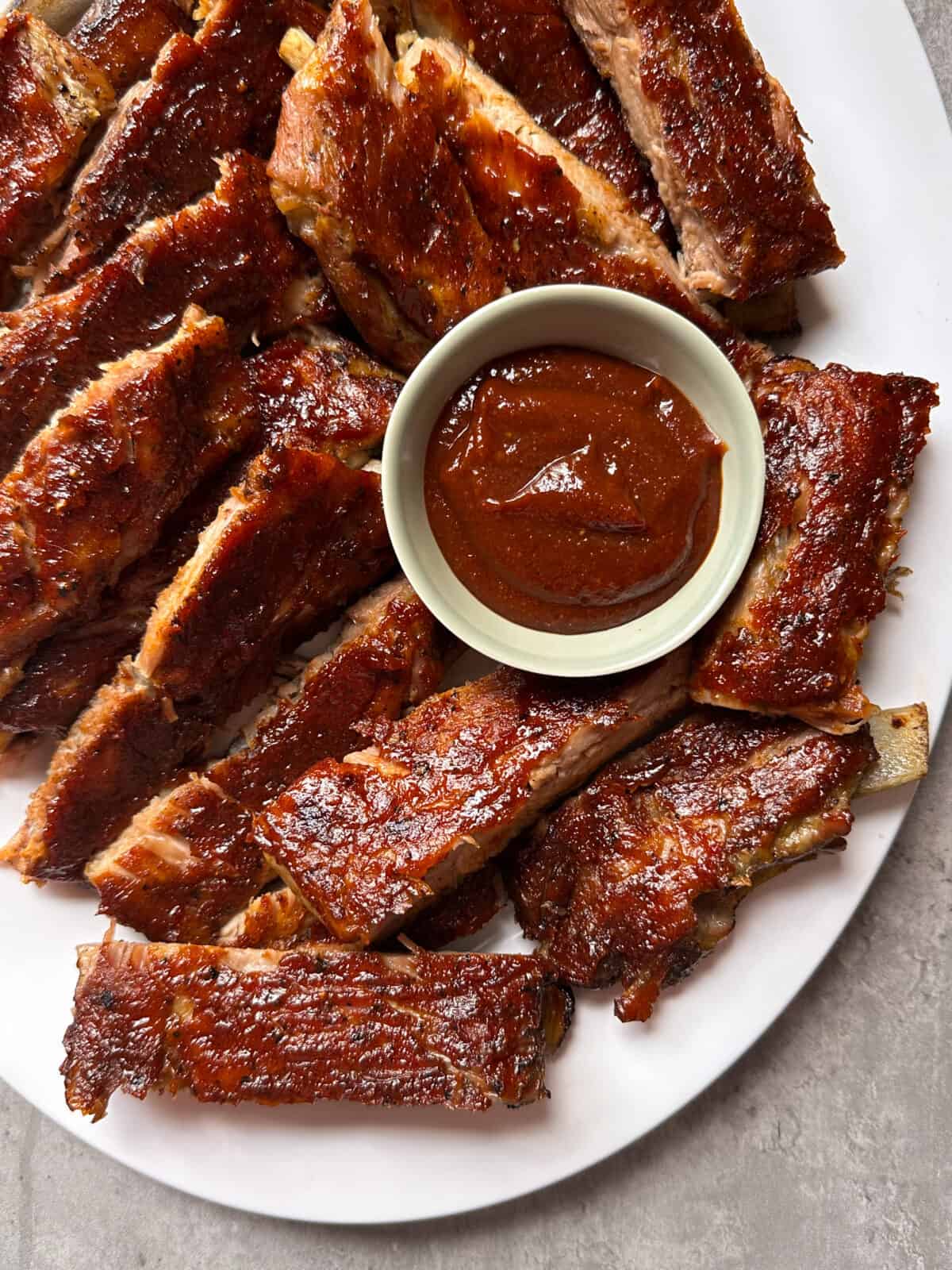 A plate of oven baked bbq ribs arranged around a small bowl of barbecue sauce, with the ribs cut into individual pieces on a white surface.
