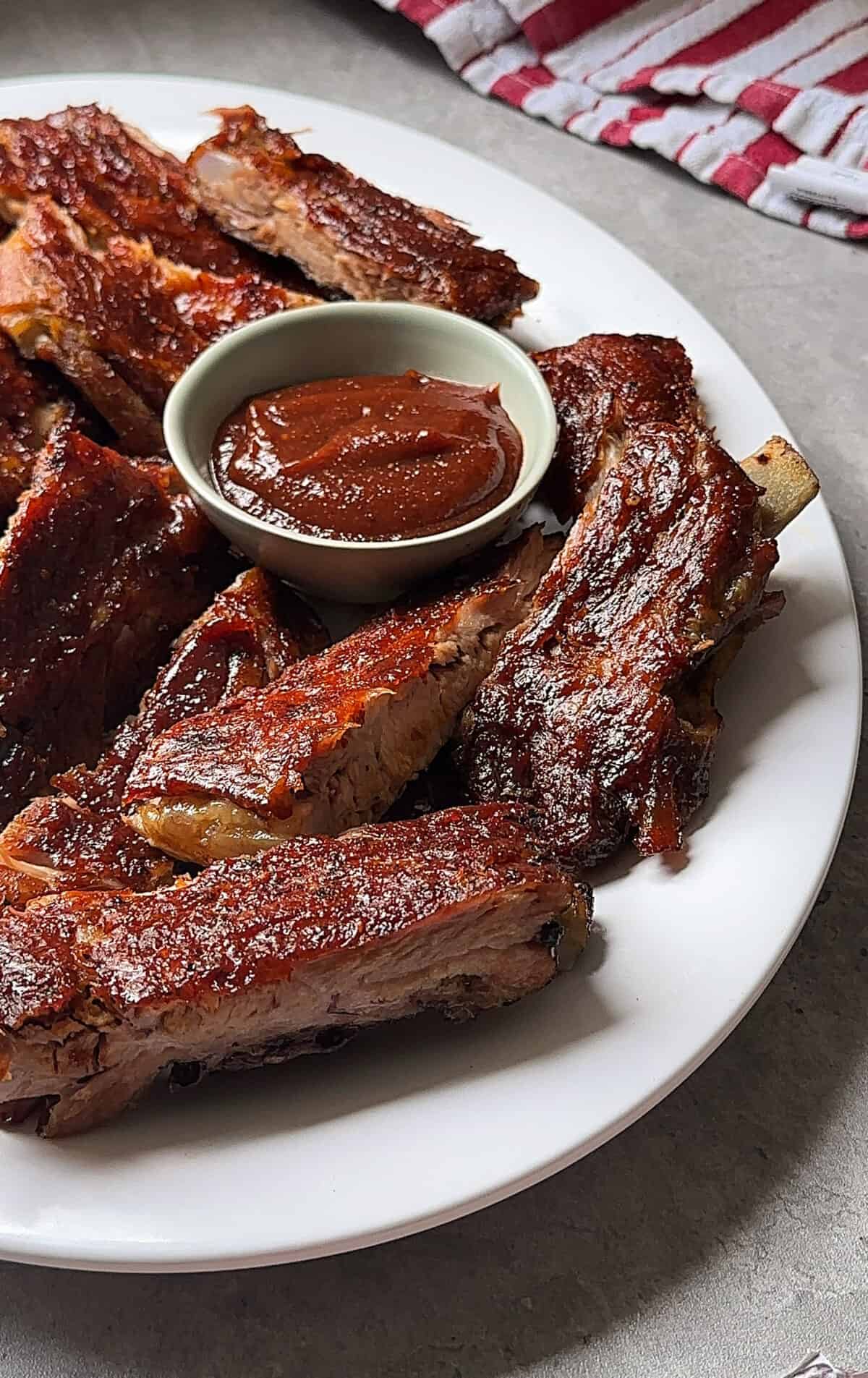 A white plate filled with oven baked bbq ribs and a small bowl of barbecue sauce, set on a gray surface with a red and white striped cloth in the background.