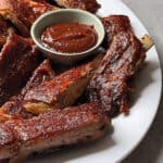 A white plate filled with juicy Oven Baked Ribs, accompanied by a small bowl of barbecue sauce. A red and white striped towel is partially visible in the background.